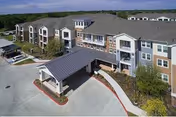Exterior view of a multi-story senior living facility building with a covered entrance driveway, surrounded by trees and parking areas under a clear sky.