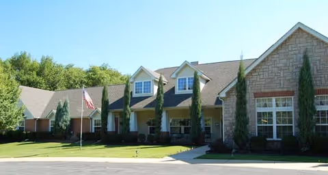 Exterior view of a single-story senior living facility building with a sloped roof, multiple windows, and a front porch area. There are tall, narrow trees and shrubs planted along the front, a flagpole with an American flag on a grassy lawn, and a paved driveway or parking area in front.