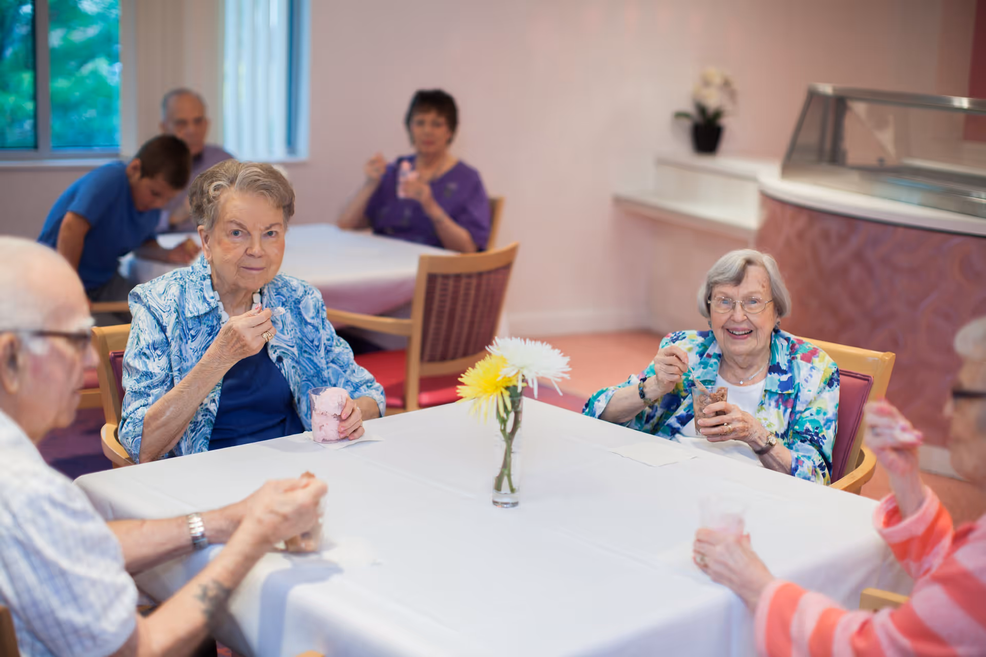 A group of elderly people sitting around a table in a dining area, enjoying desserts. The table is covered with a white tablecloth and has a small vase with flowers in the center. The room has light-colored walls and a window with vertical blinds.