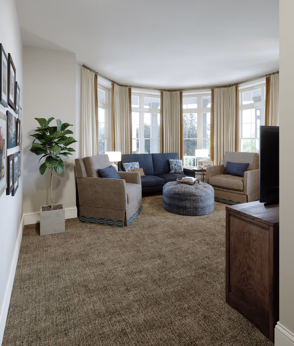 A cozy living room with beige carpet, two beige armchairs, a dark blue sofa, a round blue ottoman, and a wooden TV stand with a television. Large windows with cream-colored curtains allow natural light to fill the room. A potted plant and framed pictures decorate the space.