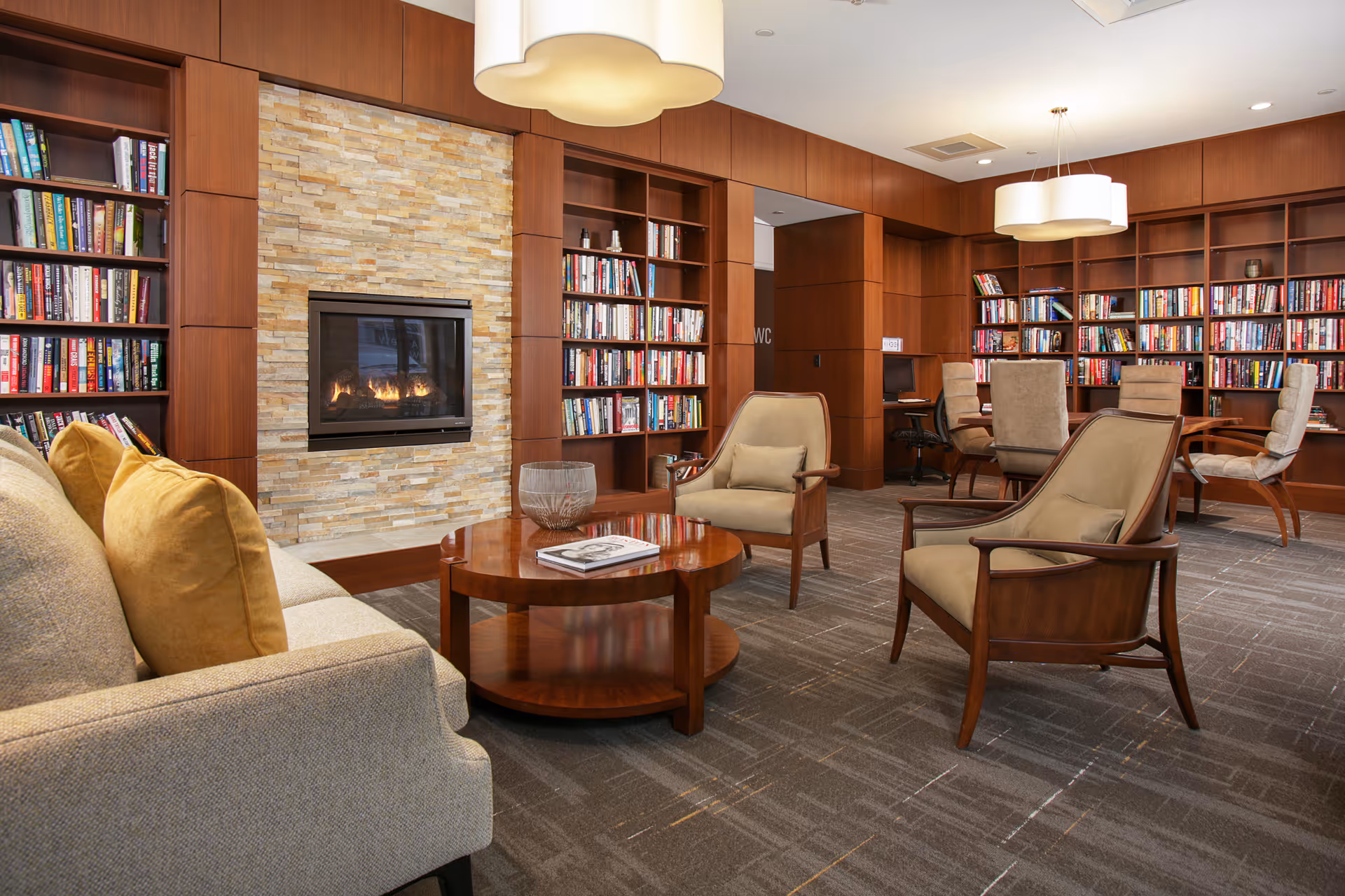 A cozy library room with wooden bookshelves filled with books, a stone fireplace with a fire burning, a beige sofa with yellow cushions, two beige armchairs, a round wooden coffee table, and a few chairs and desks in the background. The room has warm lighting and a carpeted floor.