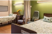 A senior living facility bedroom with two single beds, each with floral bedspreads and pillows. Between the beds is a wooden nightstand with a vase of sunflowers and a chair. A green upholstered armchair is positioned near the beds, and a curtain divider is partially drawn between the beds. The walls are painted green and beige, and there is a wall-mounted light fixture above the nightstand.