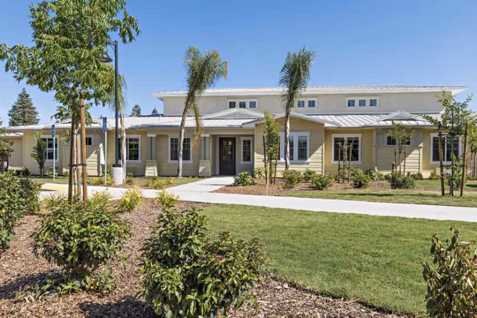 Exterior view of a single-story senior living facility building with beige siding, multiple windows, and a central entrance. The building is surrounded by landscaped greenery, including bushes, small trees, and palm trees, with a clear blue sky overhead.