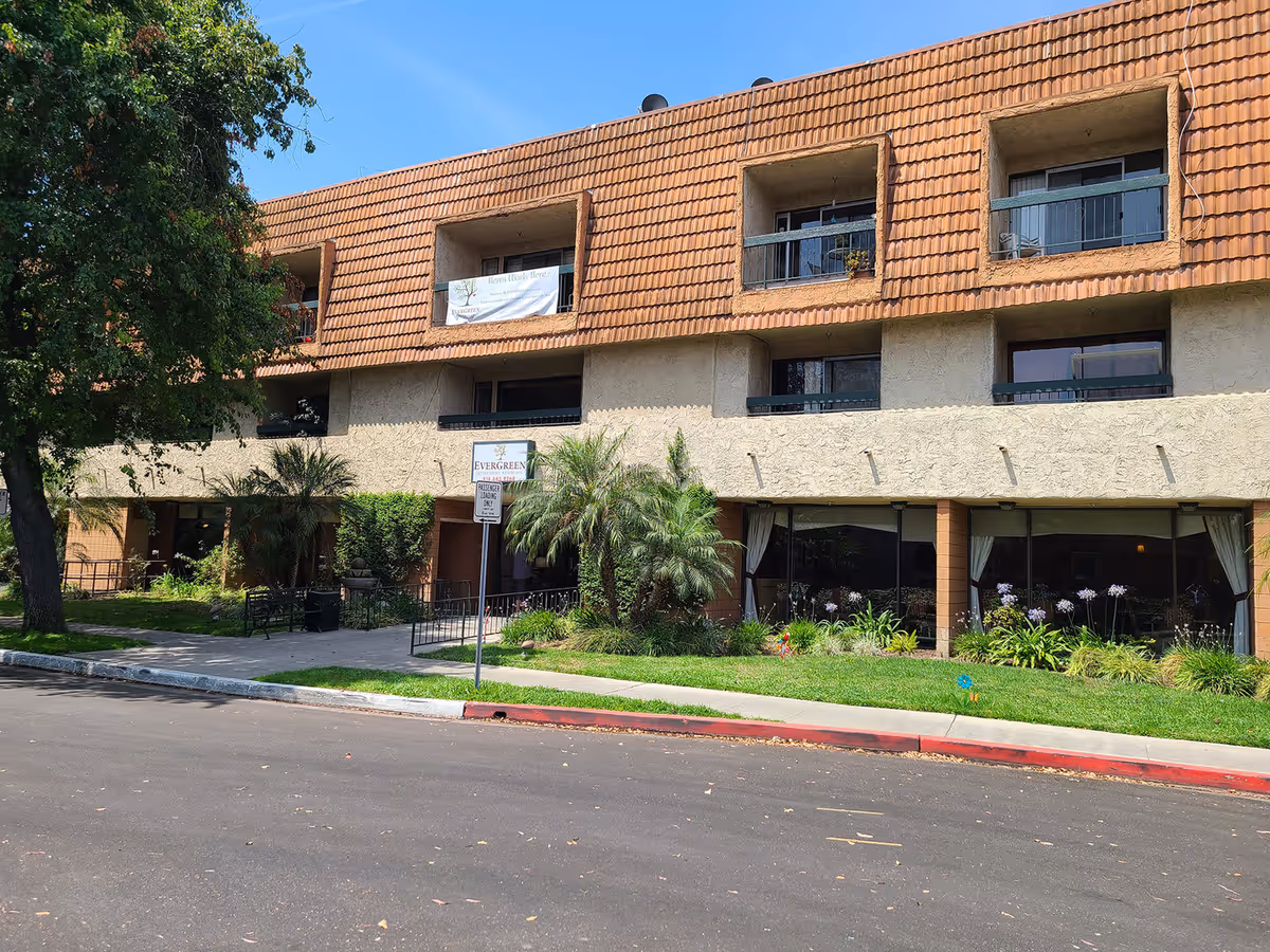 Exterior view of a three-story building with a tiled roof and balconies. The building has a sign that reads 'Evergreen Retirement Residence' near the entrance. There are trees, shrubs, and a well-maintained lawn in front of the building, along with a sidewalk and a street in the foreground.