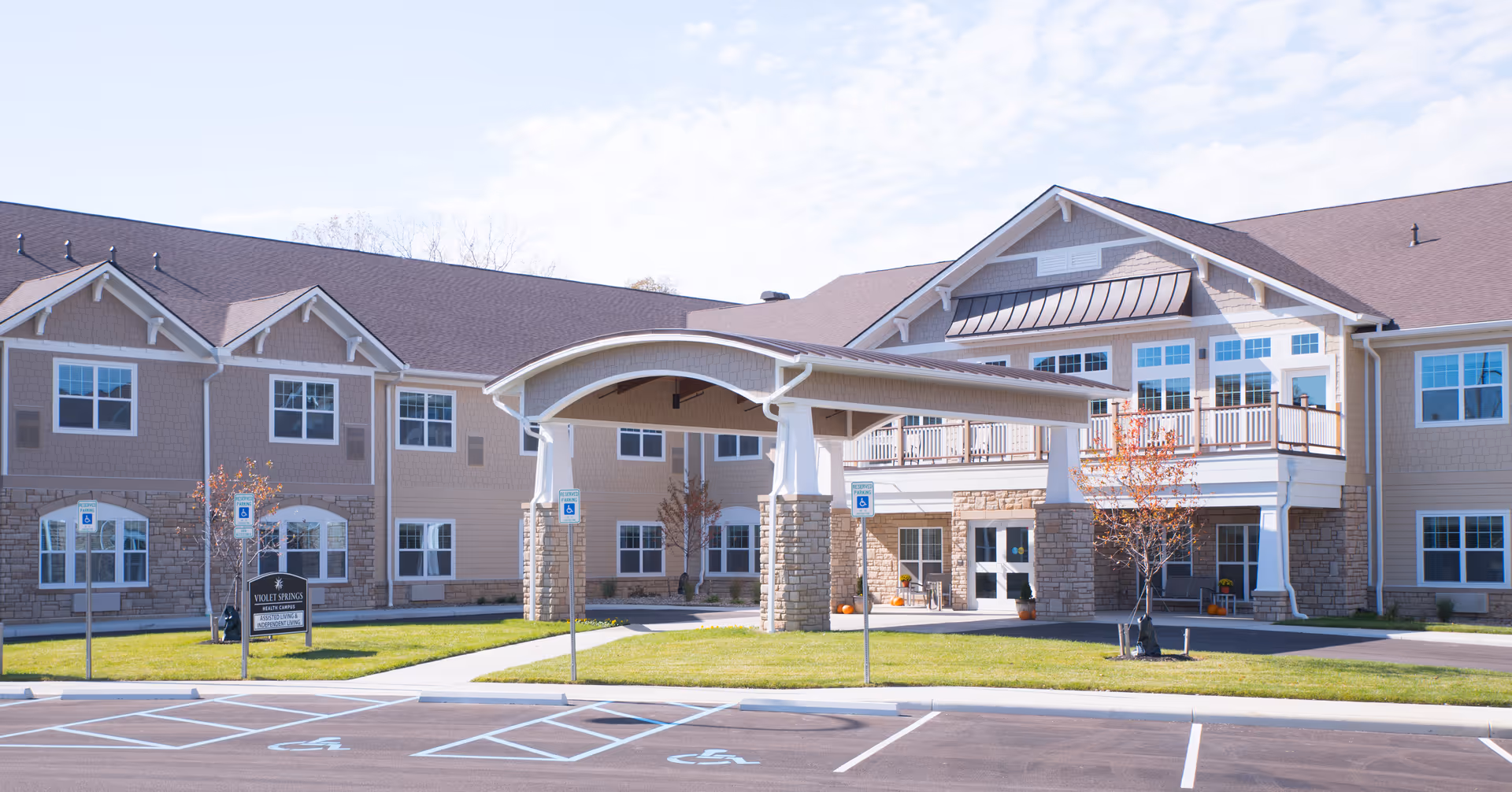 Exterior front view of Violet Springs - Senior Living Apartments building with a covered entrance supported by stone pillars, multiple windows, and a parking area with handicap spaces in front.
