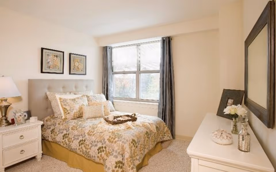 A cozy bedroom with a bed featuring floral patterned bedding and multiple pillows. There is a nightstand with a lamp and picture frames on the left side of the bed. A large window with curtains lets in natural light. On the right side, there is a dresser with decorative items and a large mirror hanging above it.
