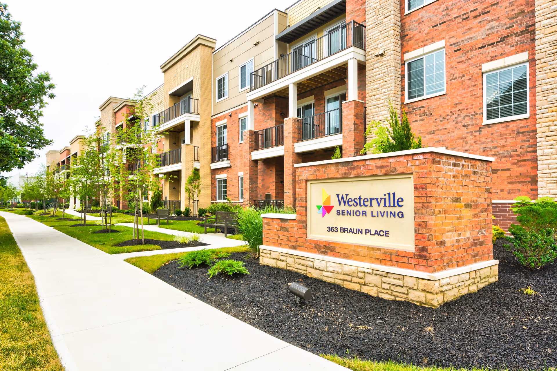 Exterior view of Westerville Senior Living facility showing a multi-story brick and stone building with balconies, a landscaped walkway with young trees and benches, and a brick sign displaying the facility name and address 363 Braun Place.