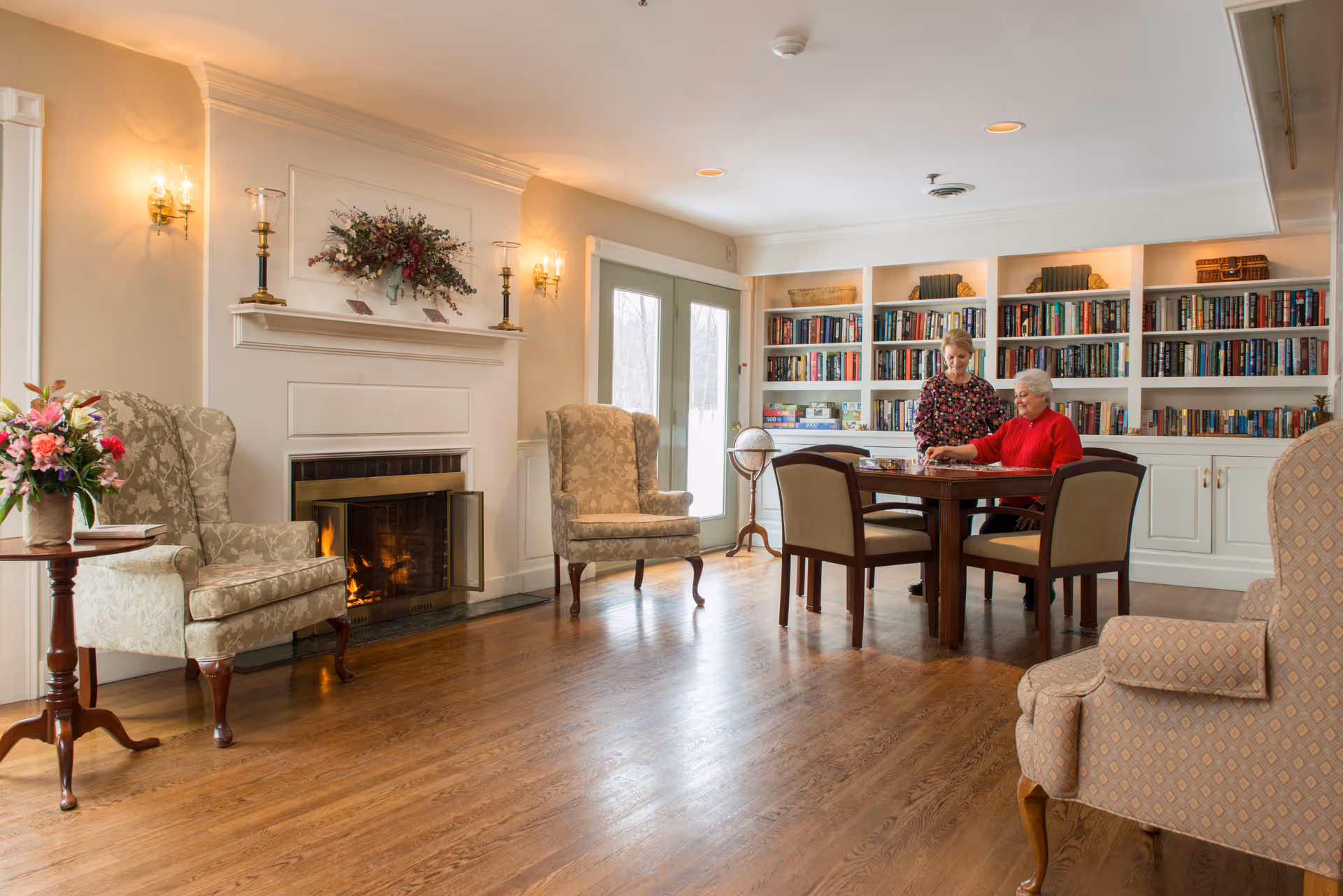 A cozy living room with wooden floors, a lit fireplace, two upholstered armchairs, a round wooden side table with a flower arrangement, and a large bookshelf filled with books. Two women are sitting at a wooden table playing a board game near the bookshelf and double glass doors.