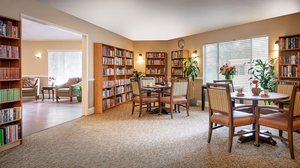 A cozy library and sitting area in a senior living facility with wooden bookshelves filled with books, round tables with chairs, potted plants, and large windows with blinds letting in natural light. In the adjacent room, there are two armchairs with a small table between them.