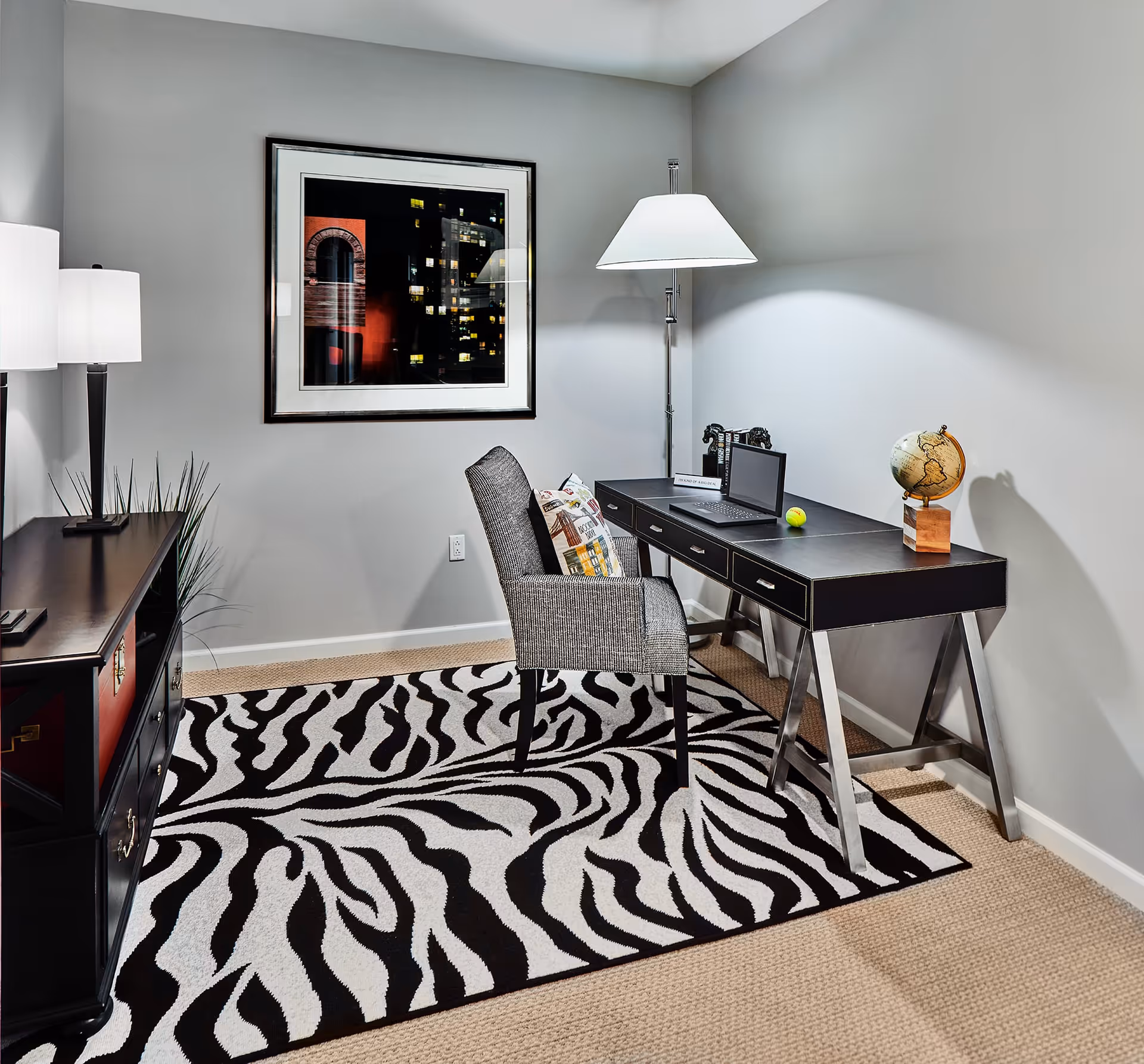 A modern home office setup with a black desk featuring three drawers, a gray upholstered chair with a decorative pillow, and a zebra-patterned rug on a beige carpet. On the desk are a laptop, a small globe, a tennis ball, and a decorative horse figurine. The room has light gray walls, a framed picture of a cityscape at night, a tall floor lamp, and a black sideboard with two table lamps and some decorative items.