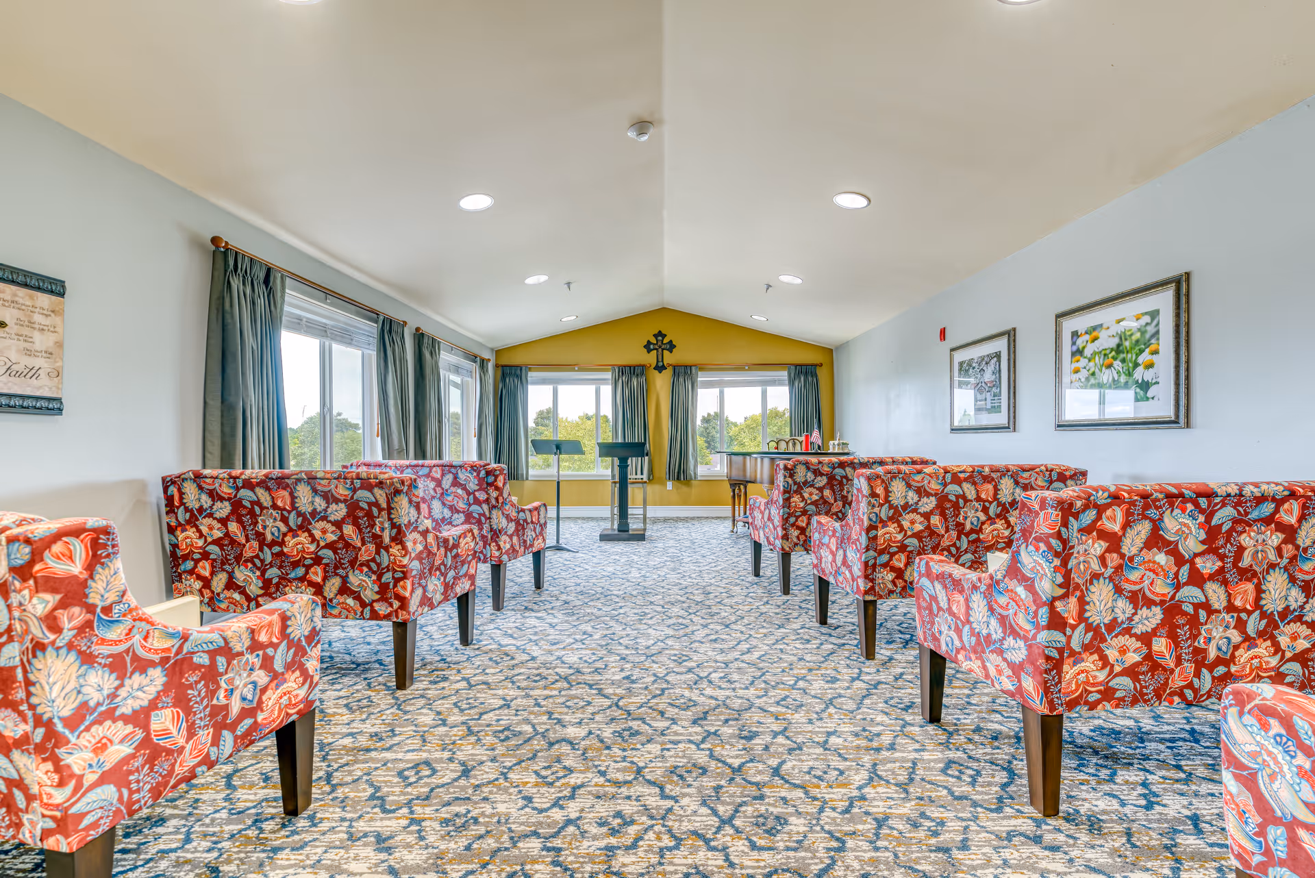 A bright room with patterned carpet and multiple red floral upholstered chairs arranged in rows facing a podium and a grand piano near large windows with green curtains. The walls are light blue with framed pictures and a cross mounted on a yellow accent wall at the far end.