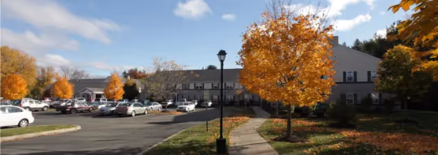 Exterior view of Carman Senior Living facility with a parking lot in front, surrounded by trees with autumn foliage under a partly cloudy sky.