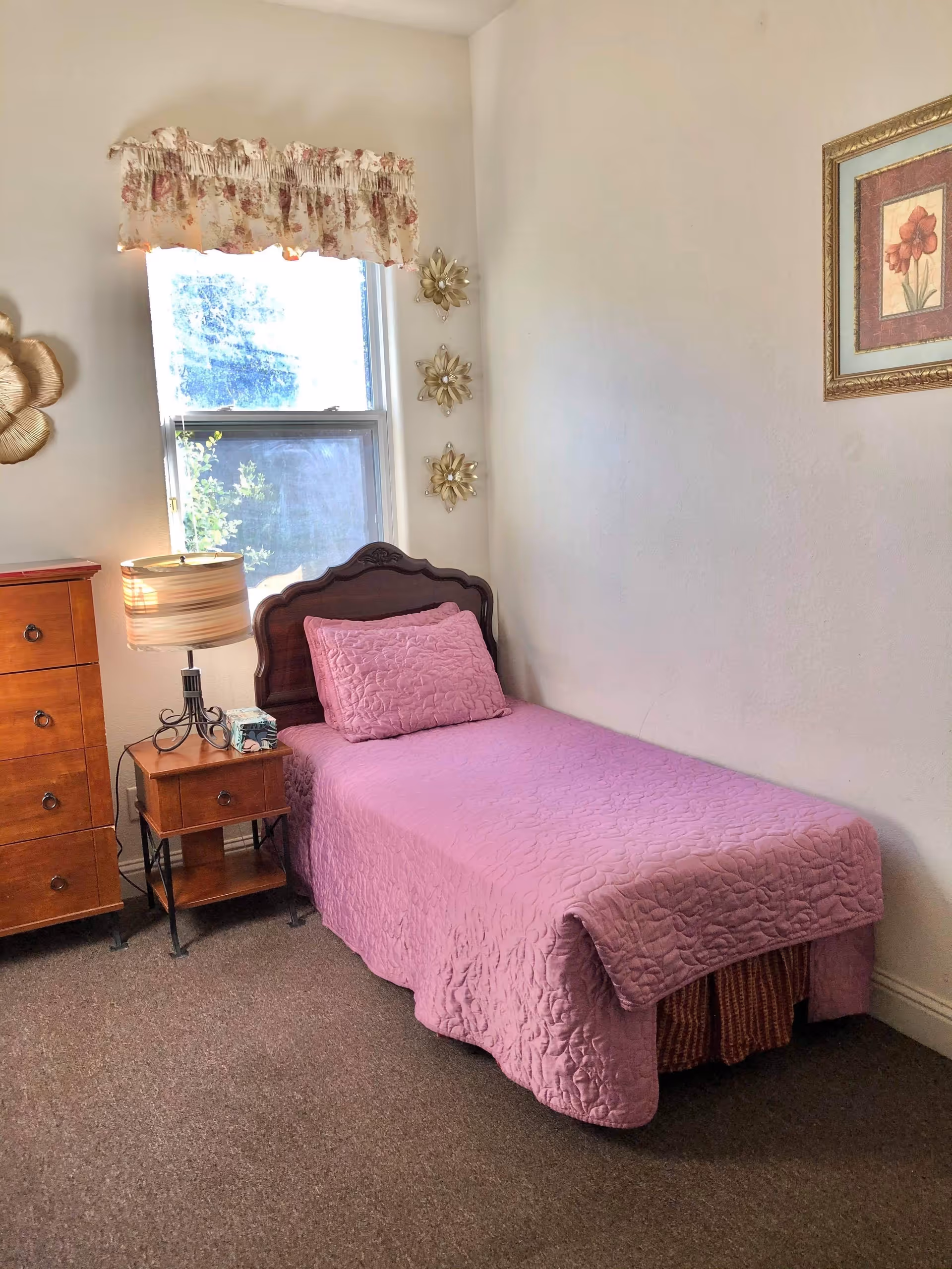 A small bedroom with a single bed covered in a pink quilt and matching pillow. Next to the bed is a wooden nightstand with a lamp and a box of tissues. There is a wooden dresser to the left of the nightstand. A window with floral valance curtains is above the nightstand, and three decorative golden wall hangings are on the wall beside the window. A framed floral picture hangs on the adjacent wall.