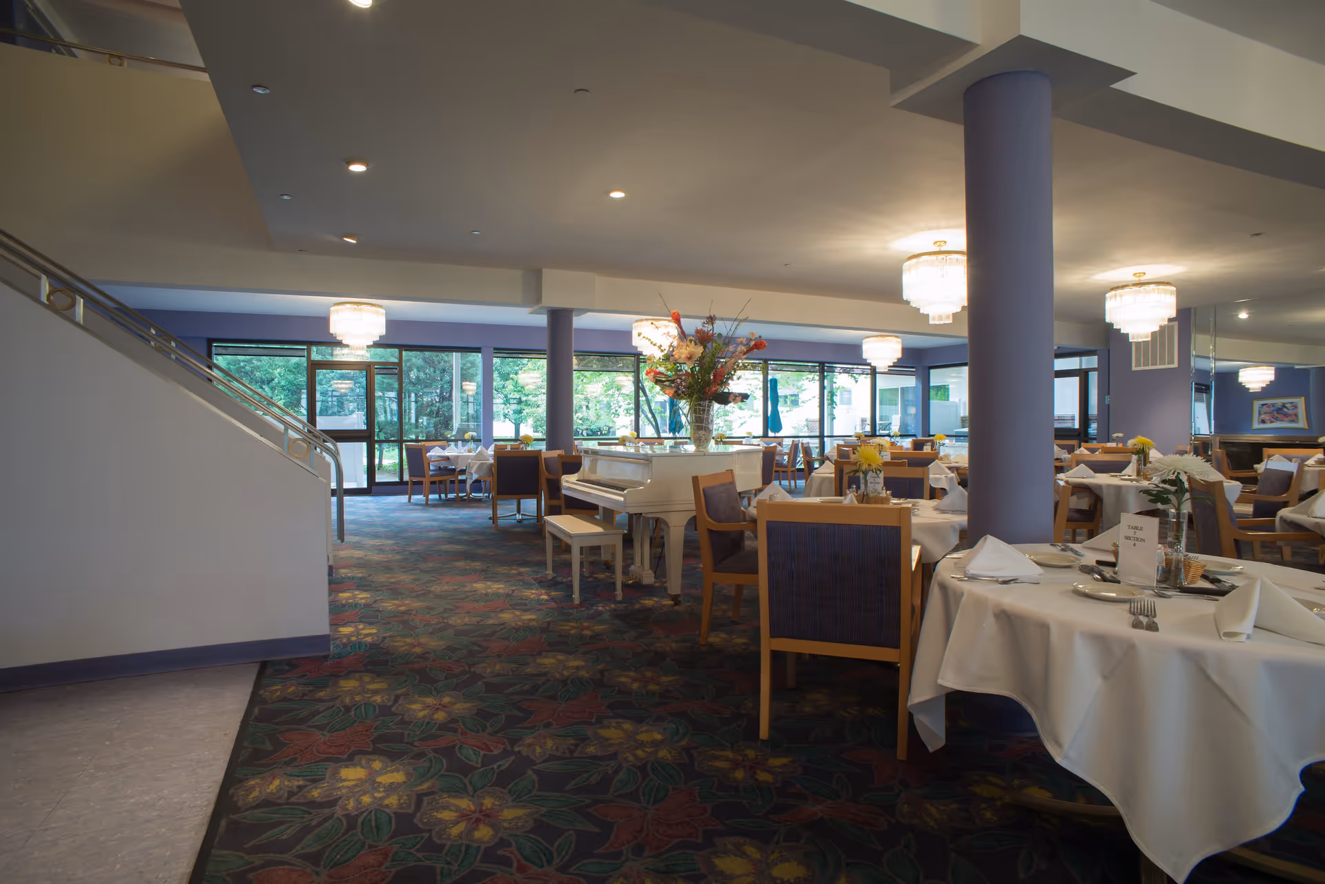 Interior view of a dining room in a senior living facility with round tables covered in white tablecloths, set with plates, napkins, and silverware. There are wooden chairs with blue cushions around the tables. A white grand piano with a floral arrangement on top is positioned near the center. Large windows and glass doors provide a view of greenery outside. The room has purple columns and ceiling lights with chandeliers.