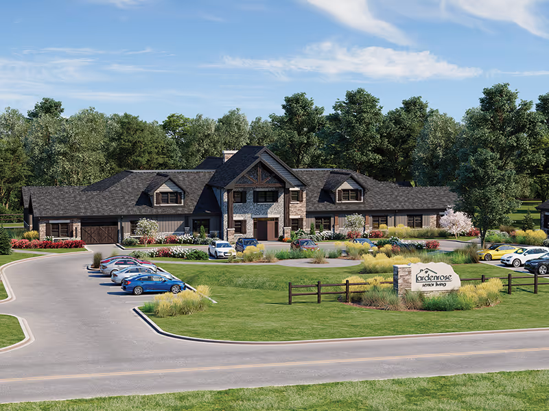 Exterior view of Arden Rose Senior Living, Inc. building surrounded by trees and landscaping with a parking lot in front containing several cars. The building has a rustic design with stone and wood elements under a blue sky with some clouds.