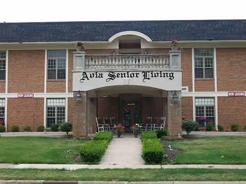 Front exterior view of a two-story brick senior living facility with a covered entrance. The building has a sign that reads 'Avia Senior Living' above the entrance, with potted plants and rocking chairs on the porch. There are small bushes and a well-maintained lawn leading up to the entrance.