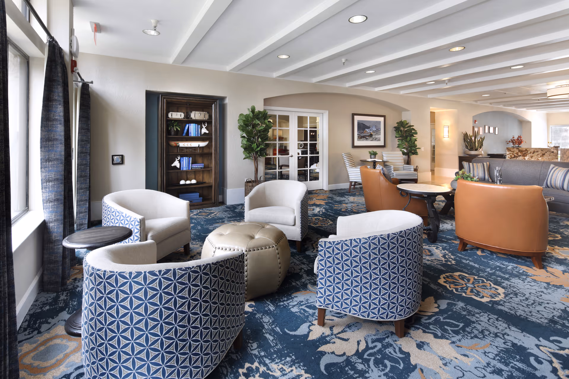 A spacious and well-lit living room area in a senior living facility featuring a variety of comfortable chairs arranged around small tables on a patterned blue carpet. The room includes a bookshelf with decorative items, potted plants, and framed artwork on the walls. Large windows with curtains allow natural light to fill the space.