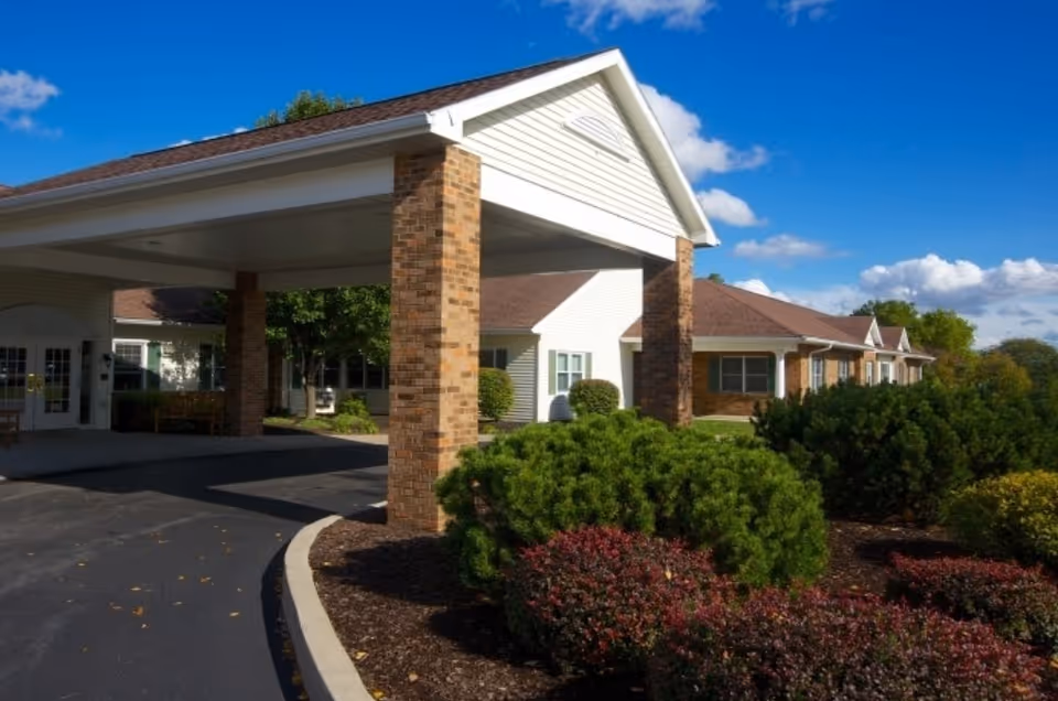 Exterior view of Keepsake Village at Greenpoint showing the entrance with a covered driveway supported by brick pillars, surrounded by well-maintained bushes and landscaping under a blue sky with some clouds.