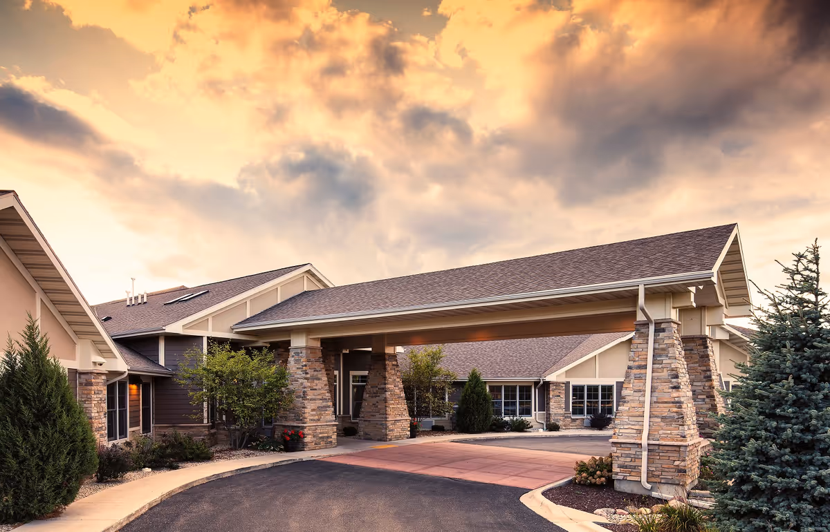 Exterior view of a senior living facility building with a covered entrance supported by stone pillars, surrounded by trees and shrubs under a dramatic cloudy sky at sunset.