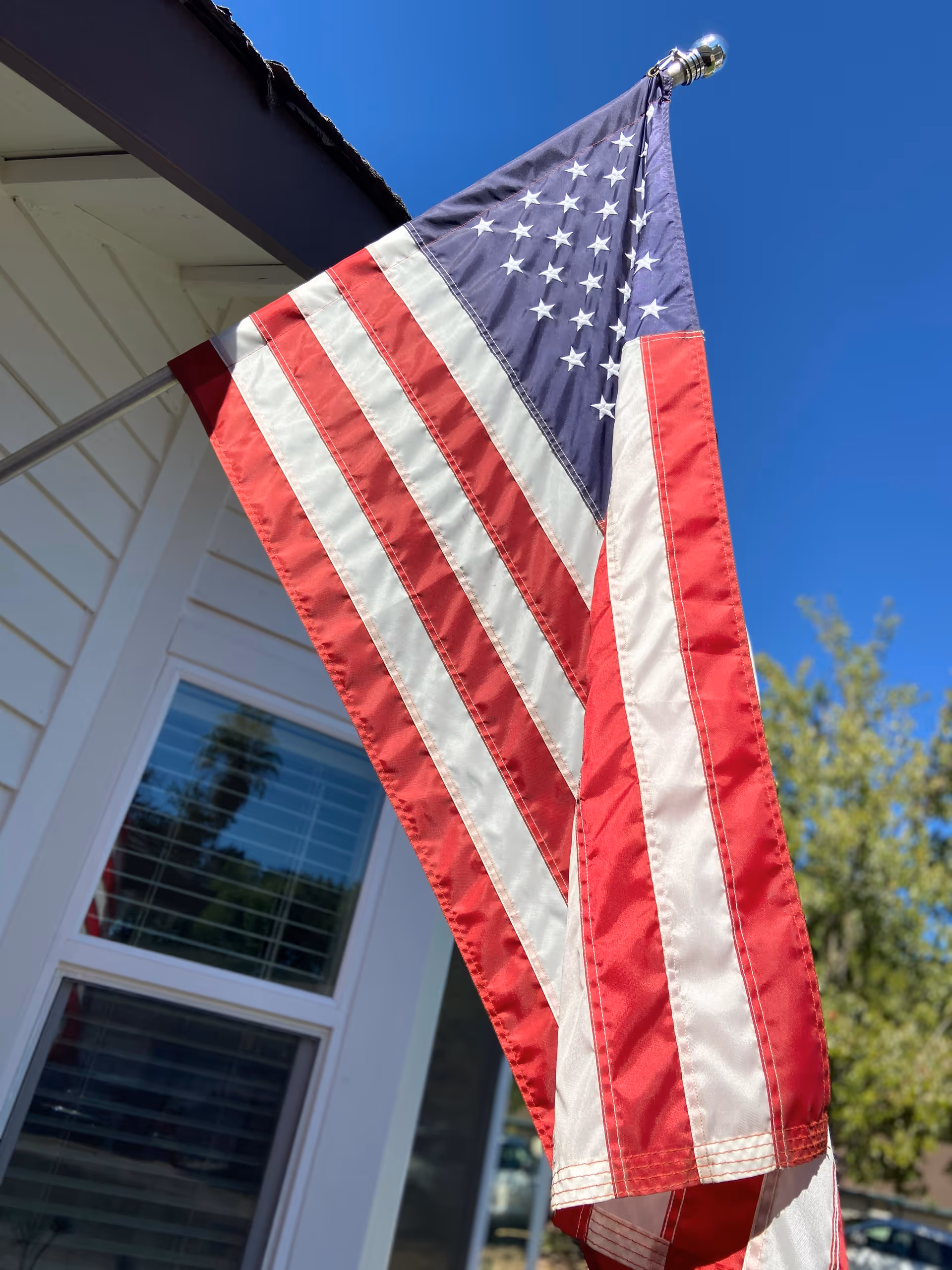 An American flag mounted on a pole attached to the exterior of a white building with a window and blue sky in the background.