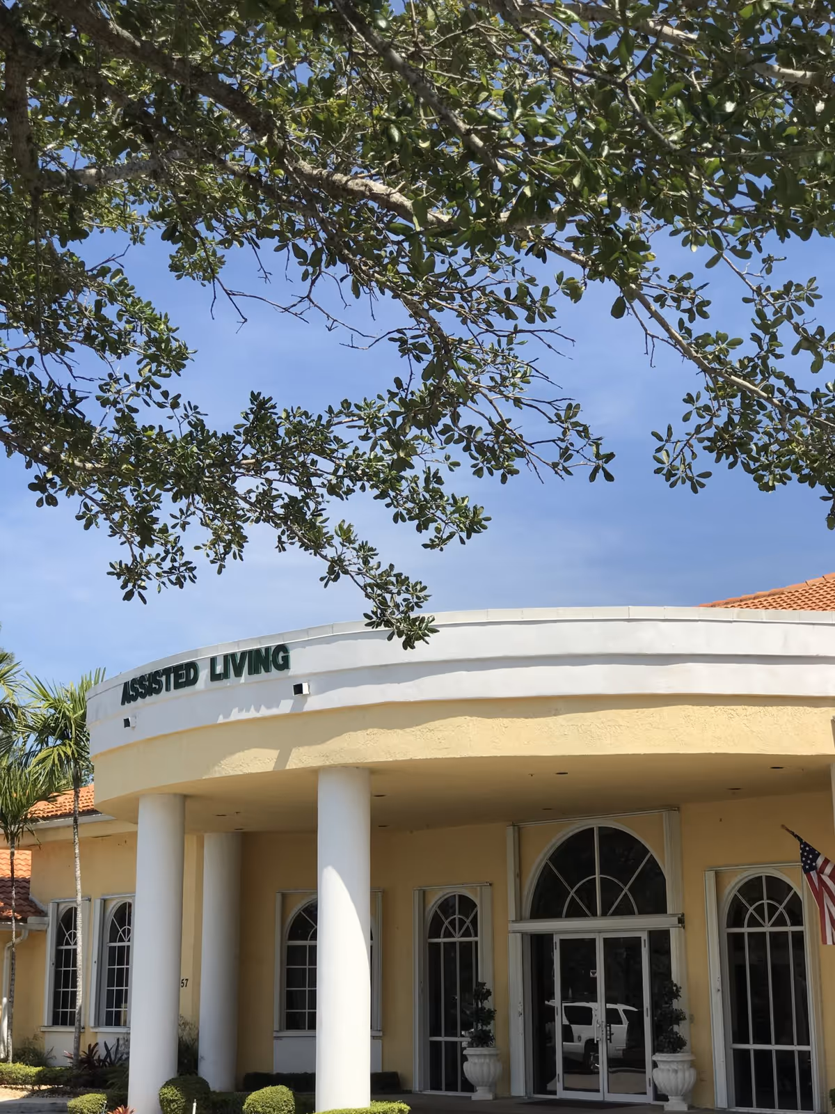Exterior view of a yellow assisted living facility building with white columns at the entrance, arched windows, and an American flag on the right side. Tree branches with green leaves are visible in the foreground against a clear blue sky.