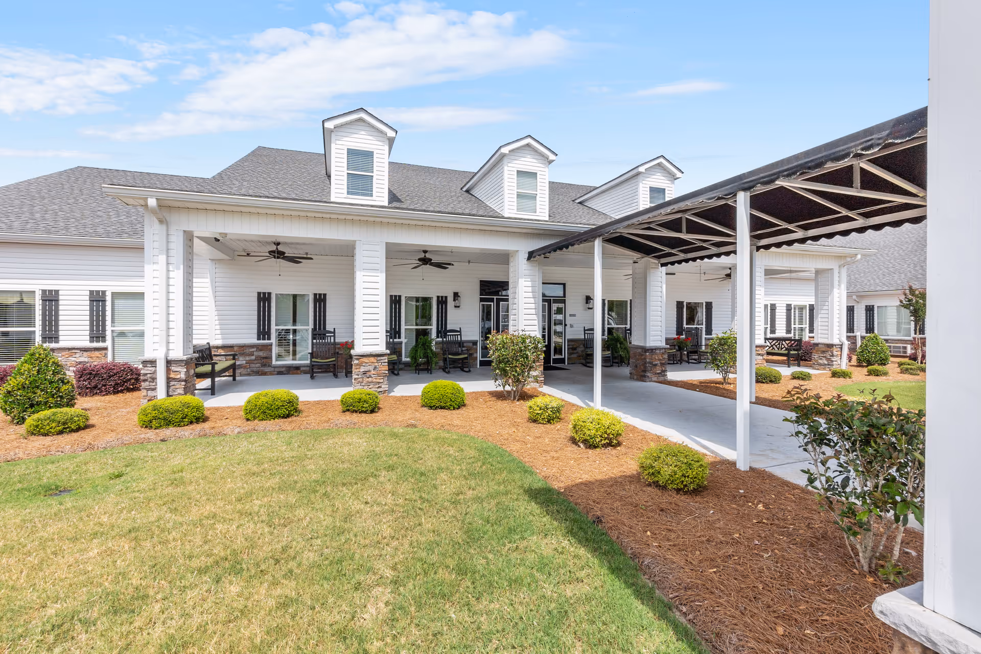 Exterior view of a senior living facility with a covered entrance, white siding, stone accents, and a well-maintained lawn with shrubs and small bushes under a blue sky.