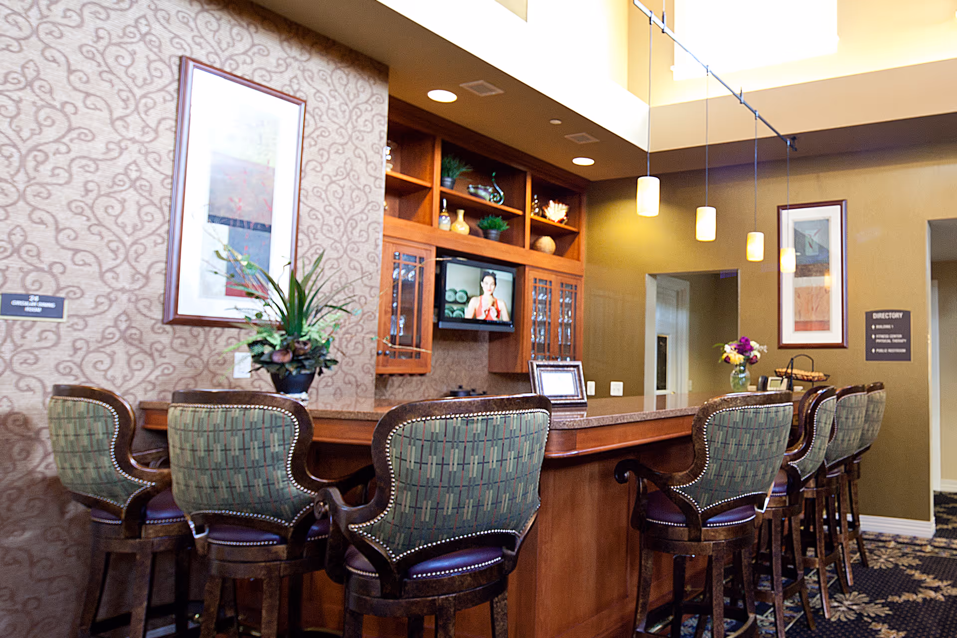 Interior view of a reception or common area in a retirement community featuring a long wooden counter with six upholstered bar stools. Behind the counter is a built-in wooden cabinet with decorative items and a small television. The walls have patterned wallpaper and framed artwork, with pendant lights hanging from the ceiling.