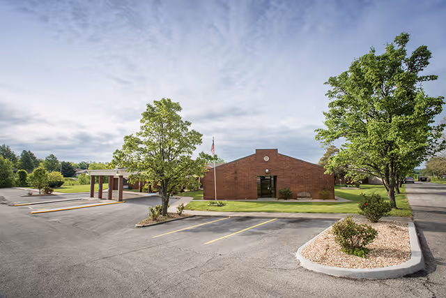 Exterior view of Pleasant Hill Health and Rehabilitation Center, a single-story brick building with a small parking lot in front, surrounded by green trees and landscaping under a partly cloudy sky.
