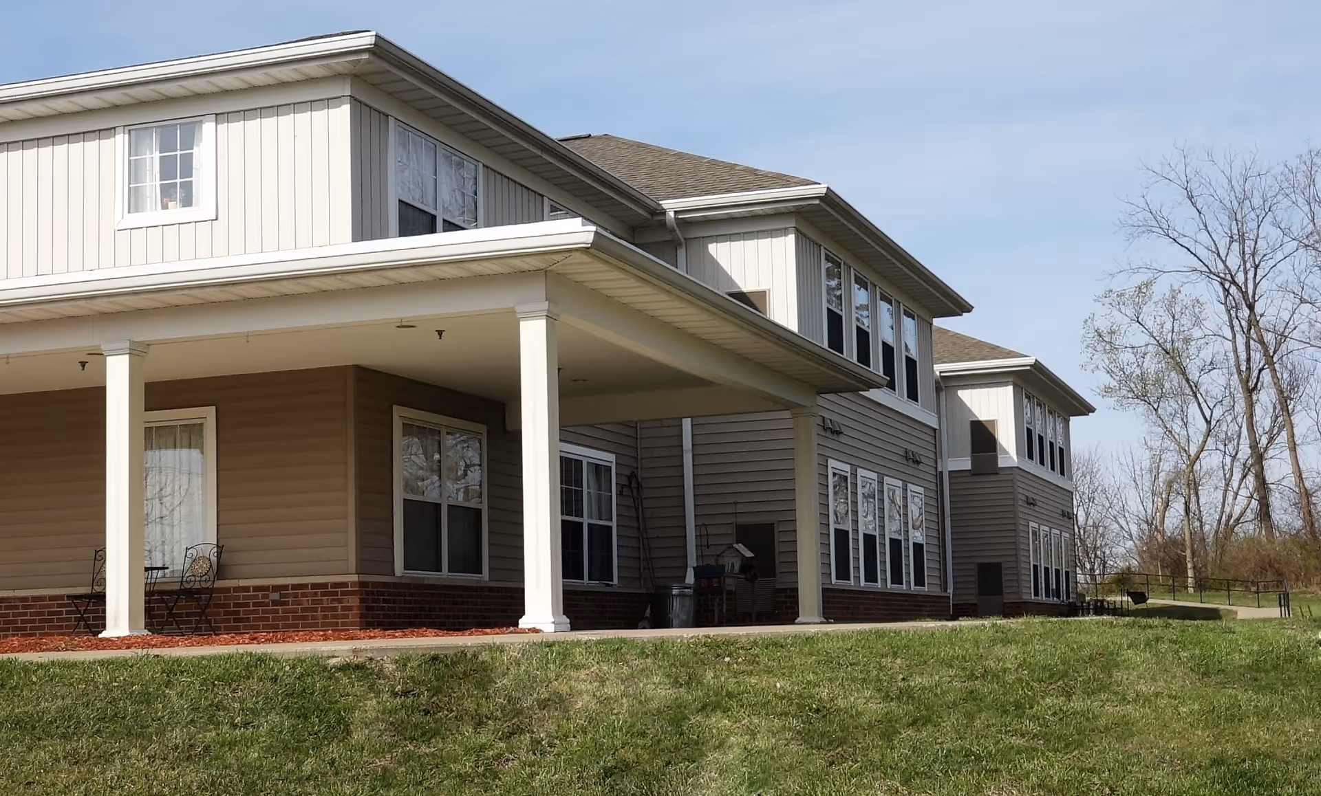 Exterior view of a two-story assisted living facility building with beige siding and white trim. The building has multiple windows and a covered porch area with white columns. There is a grassy area in the foreground and leafless trees in the background under a clear sky.