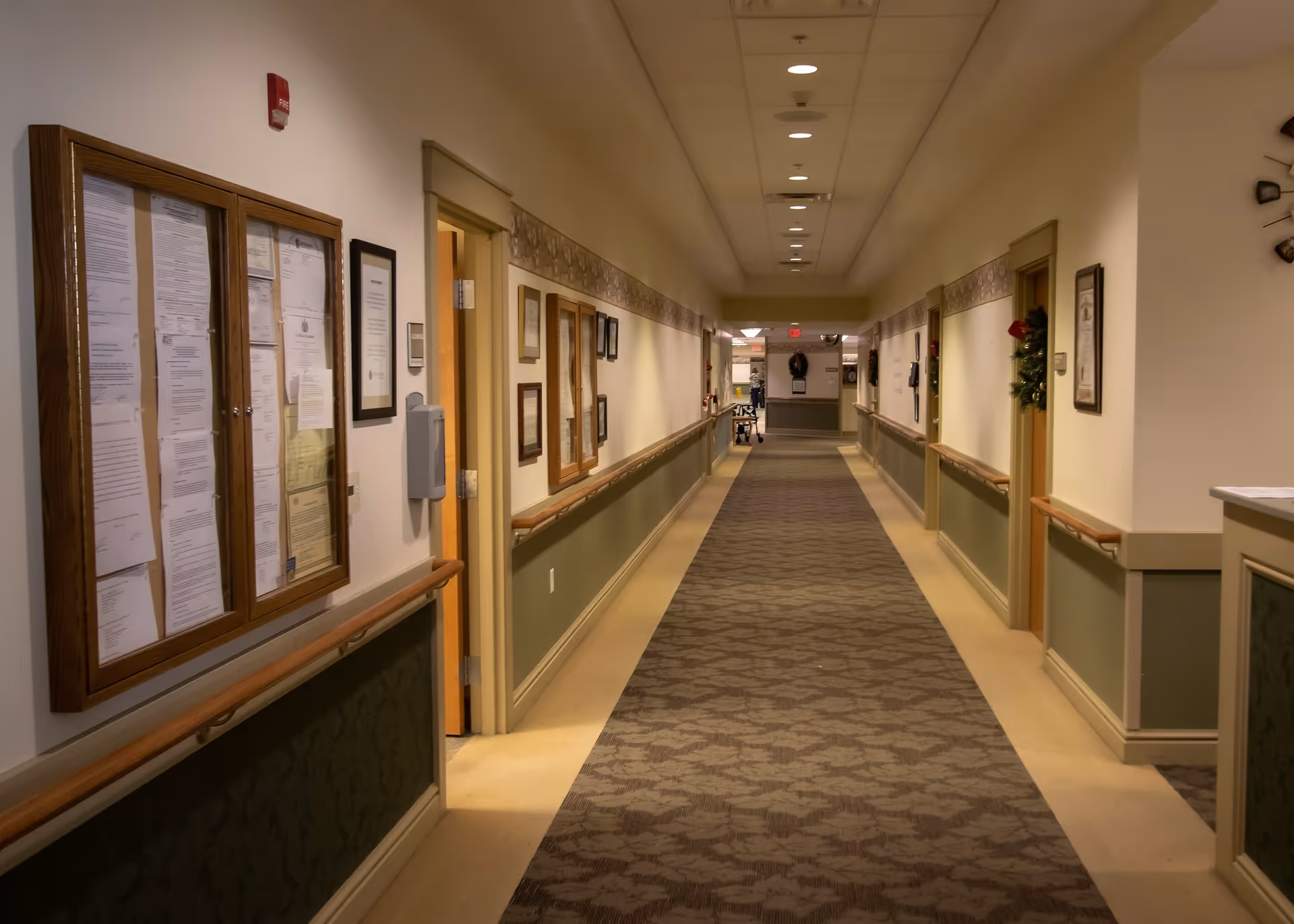 A long, well-lit hallway in a senior living facility with handrails on both sides, bulletin boards and framed documents on the walls, and several doorways leading to rooms. The floor is carpeted with a patterned runner, and there are decorations such as wreaths on some doors.