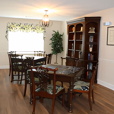 Interior view of a senior living facility dining area with wooden tables and chairs, a large window with floral curtains, a chandelier, a tall wooden bookshelf filled with books and decorative items, and a potted plant in the corner.