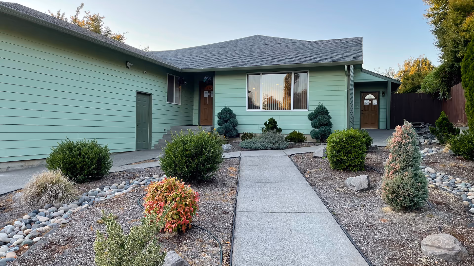 Front exterior view of a single-story building painted light green with a gray roof. There is a concrete walkway leading to two brown doors, surrounded by neatly trimmed bushes and landscaping with rocks and mulch.