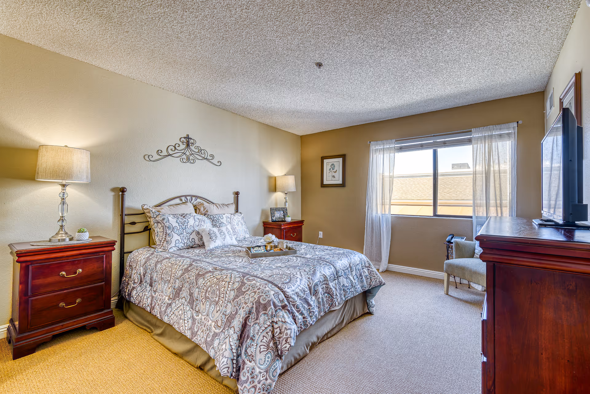 A cozy bedroom with a queen-sized bed covered in patterned bedding, two wooden nightstands with lamps on either side, a window with sheer curtains letting in natural light, a framed picture on the wall, a wooden dresser with a flat-screen TV on top, and a small upholstered chair near the window.