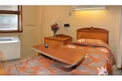 A single bed with a floral patterned bedspread in a room. There is a wooden headboard and a wooden bedside table with a vase of flowers on it. A large adjustable overbed table is positioned over the bed. A window with blinds and a wall-mounted air conditioning unit are visible.