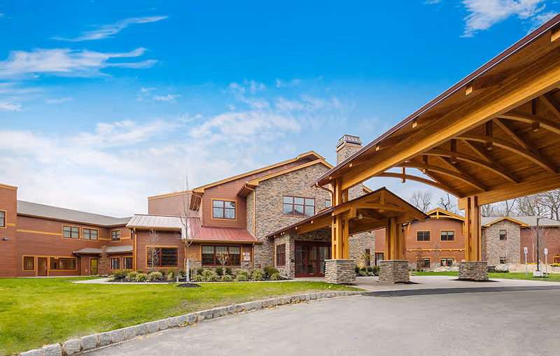 Exterior view of a large, modern rehabilitation center building with stone and wood siding, multiple windows, and a covered entrance with wooden beams. The building is surrounded by a well-maintained lawn and a paved driveway under a blue sky with some clouds.