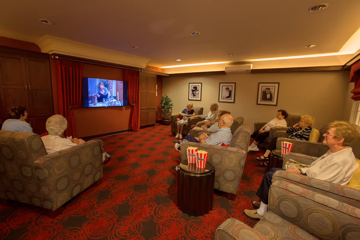A group of elderly people seated in comfortable armchairs arranged in a semi-circle in a cozy room with red patterned carpet, watching a television mounted on a wall with red curtains on either side. Small tables with popcorn containers are placed between some chairs. The walls are decorated with framed black and white portraits and there is a potted plant in the corner.