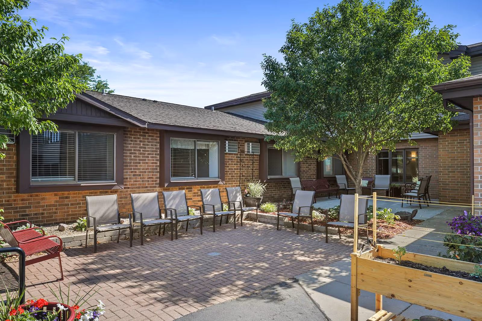 Outdoor courtyard area at Waunakee Valley Senior Living featuring a paved patio with multiple chairs arranged around the perimeter, a tree providing shade, flower beds, and a raised wooden planter box with plants. The surrounding building has brick walls and windows with blinds.