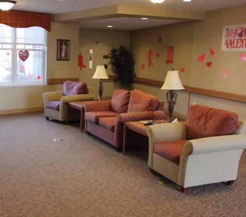 A cozy living room area in a senior living facility with three upholstered chairs and a loveseat arranged along a wall. Two table lamps sit on wooden side tables between the seating. The walls are decorated with Valentine-themed paper cutouts and a banner. A large window with a red valance allows natural light into the room.