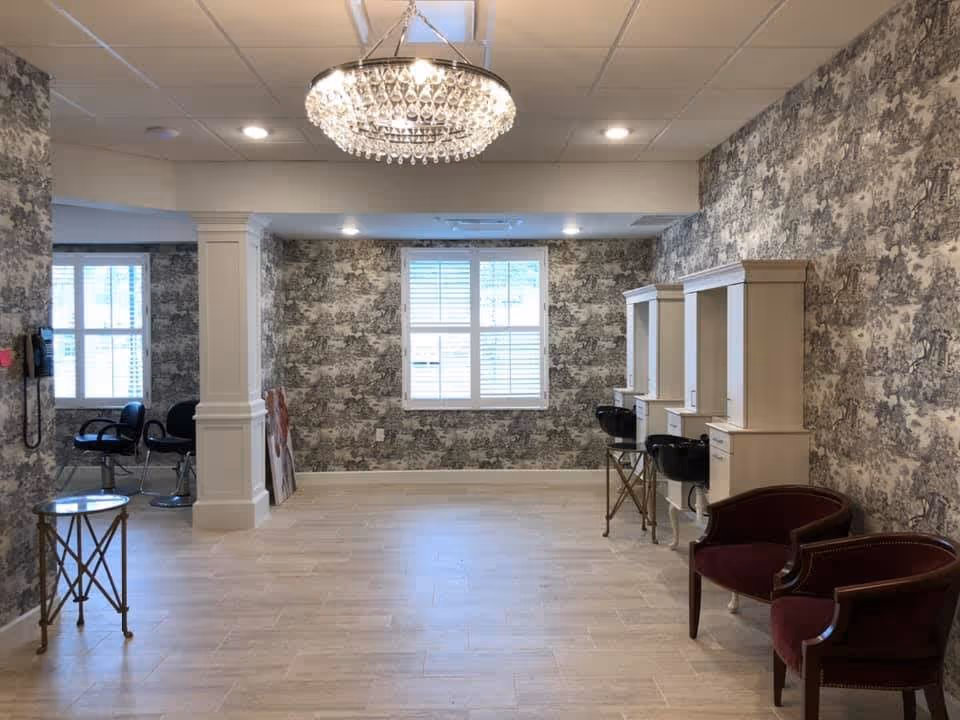 Interior room with patterned wallpaper, a chandelier, two burgundy armchairs, two white vanity stations with black sinks, and a few black chairs near windows with white shutters.