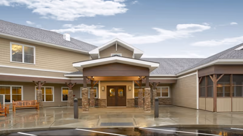 Exterior view of Valley Residential Services building entrance with a covered porch supported by stone pillars, beige siding, and multiple windows. There is a bench to the left and a screened porch area to the right under a cloudy sky.