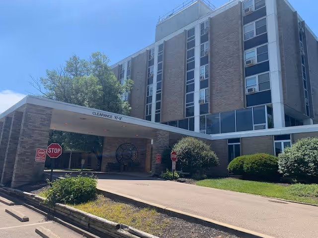 Exterior view of a multi-story brick building with large windows and a covered entrance driveway. There are stop signs near the entrance and well-maintained bushes and trees around the building under a clear blue sky.