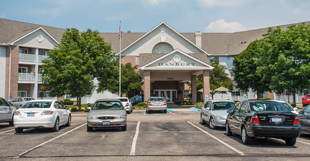 Front exterior view of Danbury North Canton facility showing a three-story building with a covered entrance, several parked cars in the parking lot, trees, and a flagpole with an American flag.