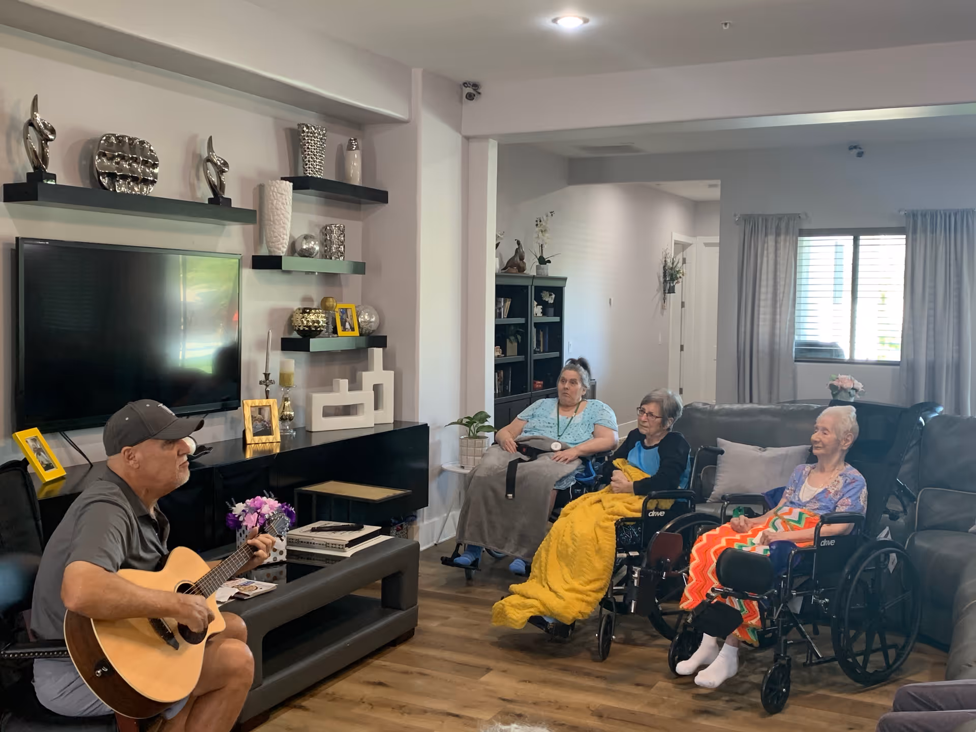 A man wearing a cap is playing an acoustic guitar in a living room area. Three elderly women, two in wheelchairs and one seated on a couch, are watching and listening. The room has a modern entertainment center with a flat-screen TV, decorative shelves with vases and picture frames, and a window with curtains letting in natural light.