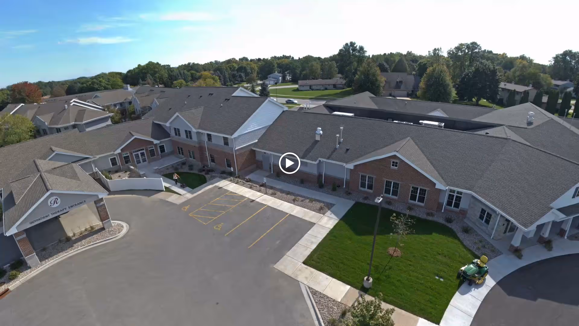 Aerial view of a senior living facility building with a large parking lot, green lawns, and surrounding trees under a blue sky. The building has multiple sections with pitched roofs and brick and siding exteriors. A sign on one section reads 'Outpatient Therapy Entrance.'