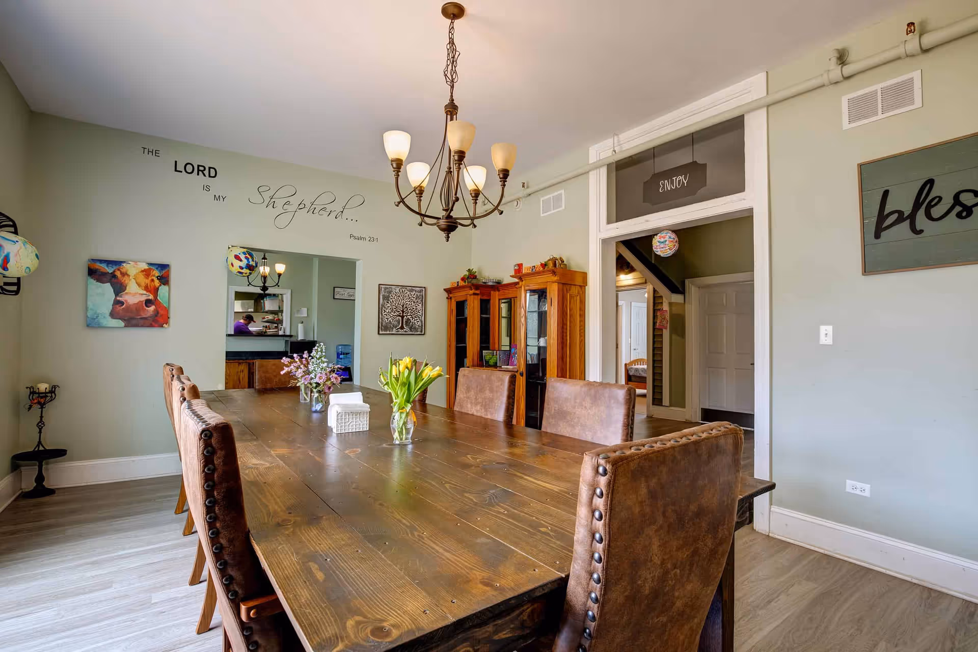 A dining room with a long wooden table surrounded by brown leather chairs. The walls are painted light green and decorated with artwork, including a painting of a cow and a tree, and wall text that reads 'The Lord is my Shepherd... Psalm 23:1'. A chandelier with six lights hangs above the table. There are two wooden cabinets against the far wall and a doorway leading to another room with a sign above it that says 'Enjoy'.