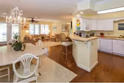 Open interior space showing a kitchen with white cabinets and countertops on the right, a dining area with a white table and chairs in the foreground, and a living room with sofas, armchairs, and a chandelier in the background. The room has hardwood floors and large windows letting in natural light.