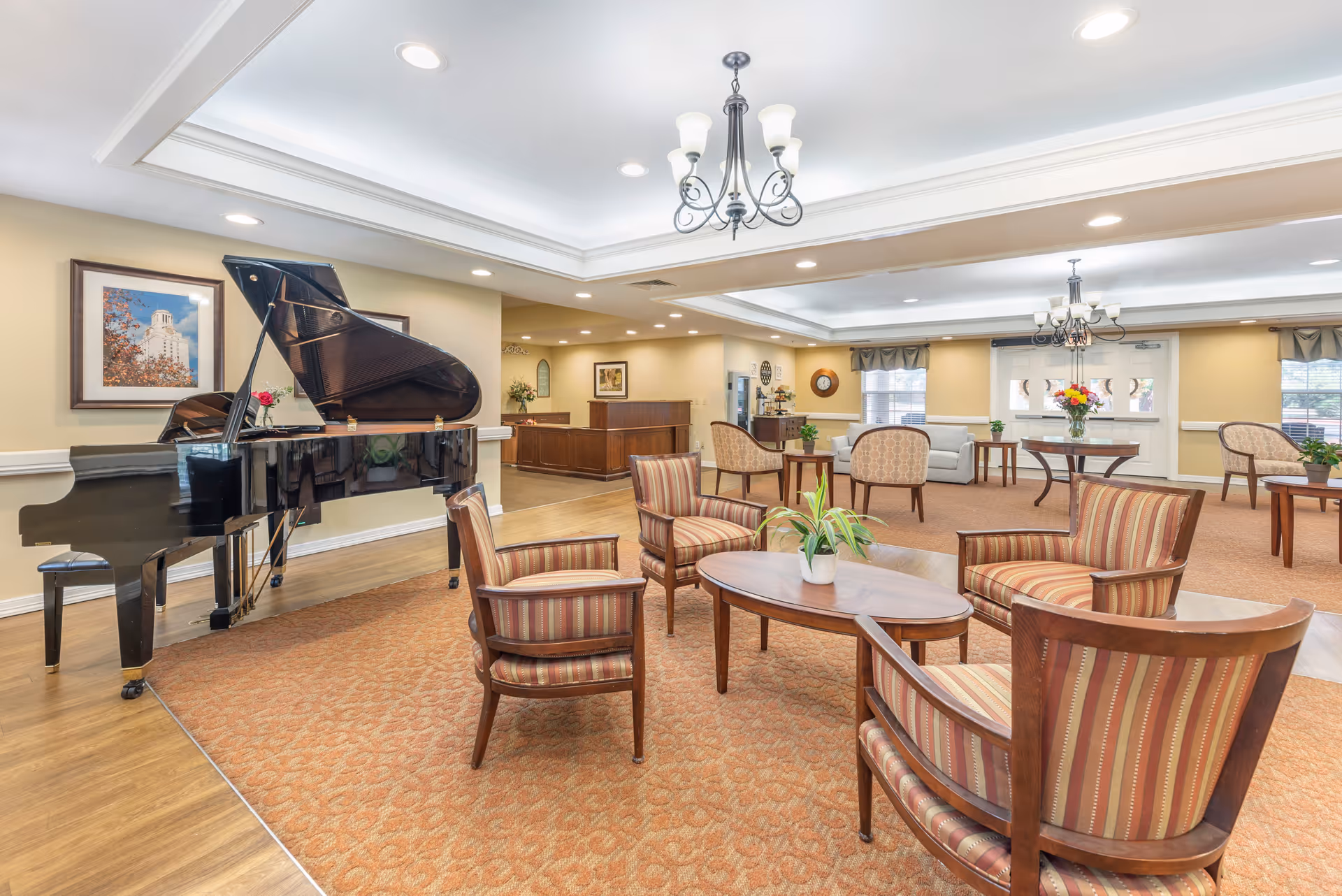 A spacious and well-lit common area in a senior living facility featuring a black grand piano, several upholstered chairs with striped fabric arranged around wooden coffee tables, and additional seating areas with armchairs and a sofa. The room has beige walls, framed artwork, chandeliers, and large windows with curtains allowing natural light to enter.