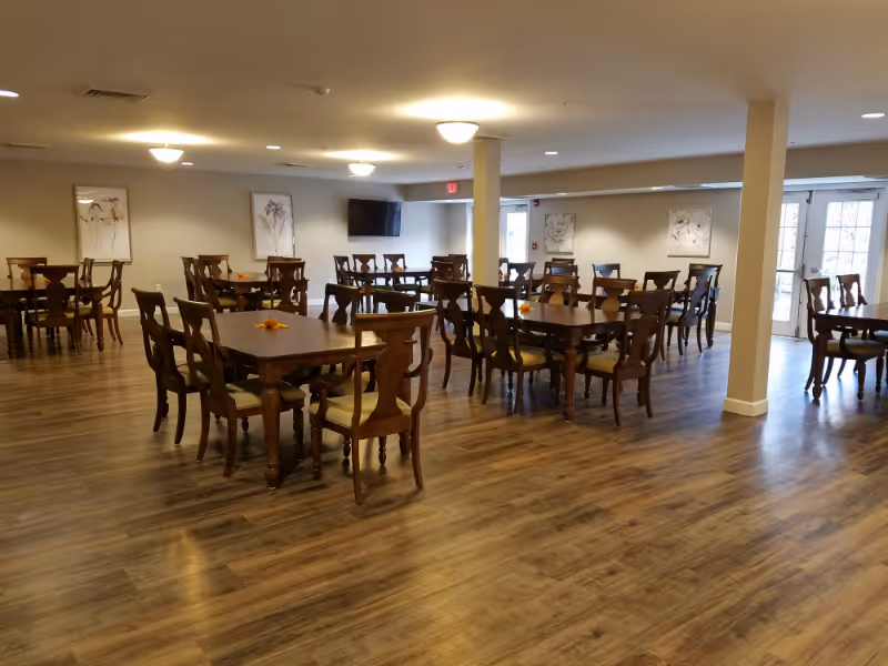 A spacious dining room with multiple wooden tables and chairs arranged neatly on a wood-patterned floor. The room is well-lit with ceiling lights and has light-colored walls adorned with framed artwork. There is a flat-screen TV mounted on one wall and large windows or glass doors allowing natural light to enter.