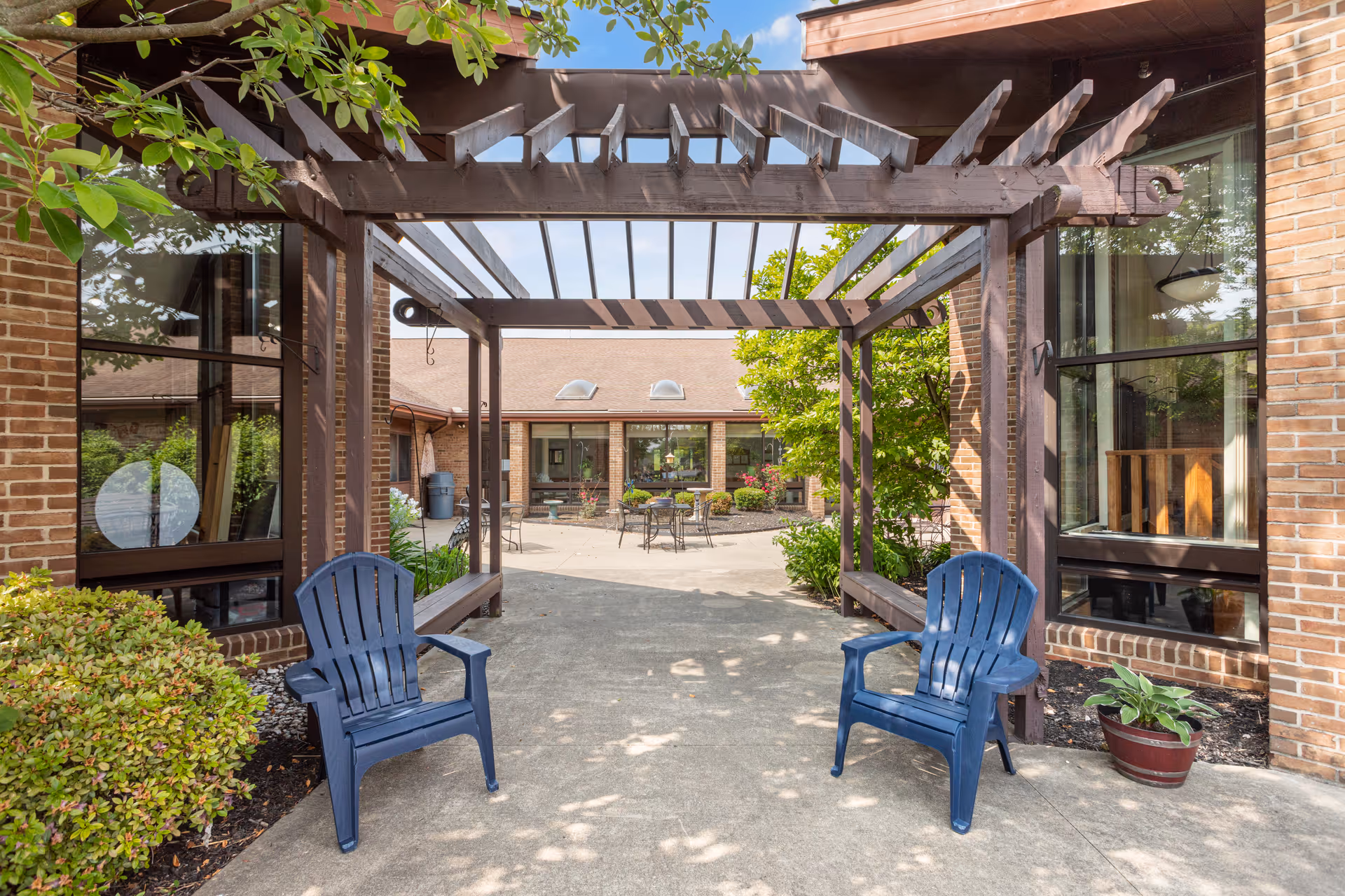 Outdoor courtyard area at Crown Pointe Care Center featuring a wooden pergola with two blue plastic chairs underneath. The courtyard is surrounded by brick buildings with large windows, greenery, and potted plants. There are tables and chairs visible further inside the courtyard under a clear sky.