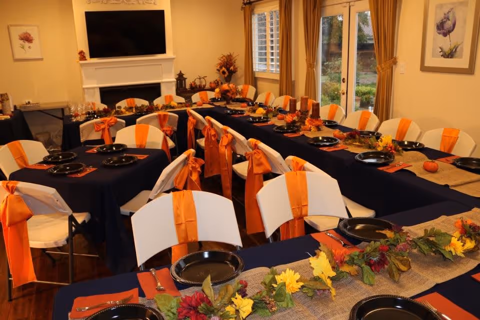 A dining room set up for a festive meal with tables covered in dark blue tablecloths and decorated with orange ribbons tied to white folding chairs. The tables have black plates, orange napkins, silverware, and autumn-themed centerpieces including leaves, flowers, and small pumpkins. The room has a fireplace with a TV mounted above it, windows with beige curtains, and framed floral artwork on the walls.